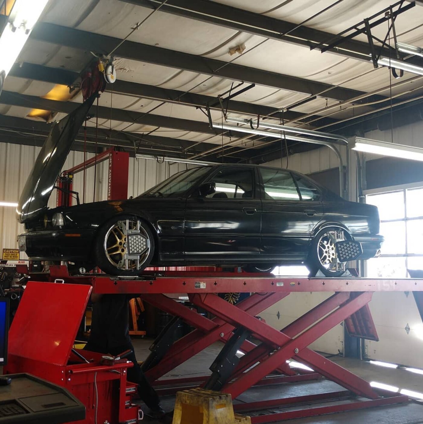A black BMW E34 raised on an alignment lift inside the shop, hood open