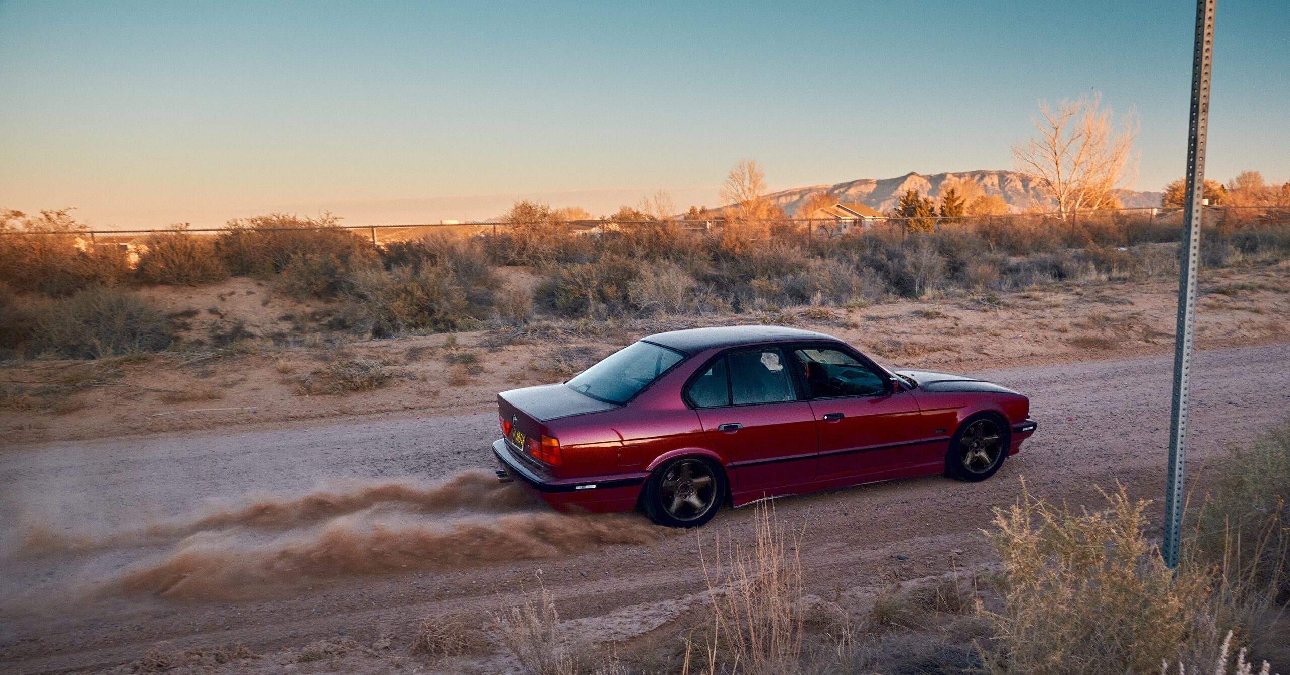 A red BMW E34 drifting on a dirt road at golden hour, with the Sandia Mountains in the background near Albuquerque, New Mexico